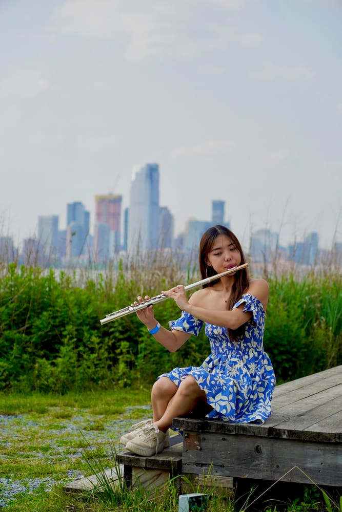 Monica playing the flute on a dock, a city skyline behind her