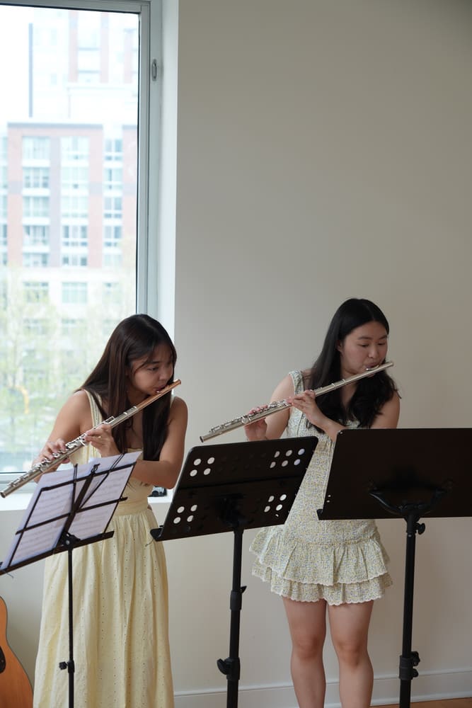 Two flutists performing at music stands by a window