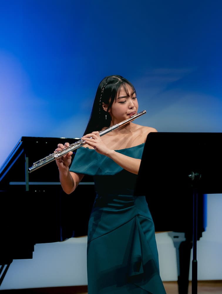 Monica playing the flute beside a grand piano under blue light